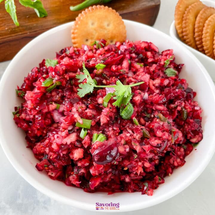 A white bowl filled with finely chopped cranberry salsa, garnished with cilantro, sits next to crackers on a white surface-a vibrant and refreshing twist for your next appetizer spread.