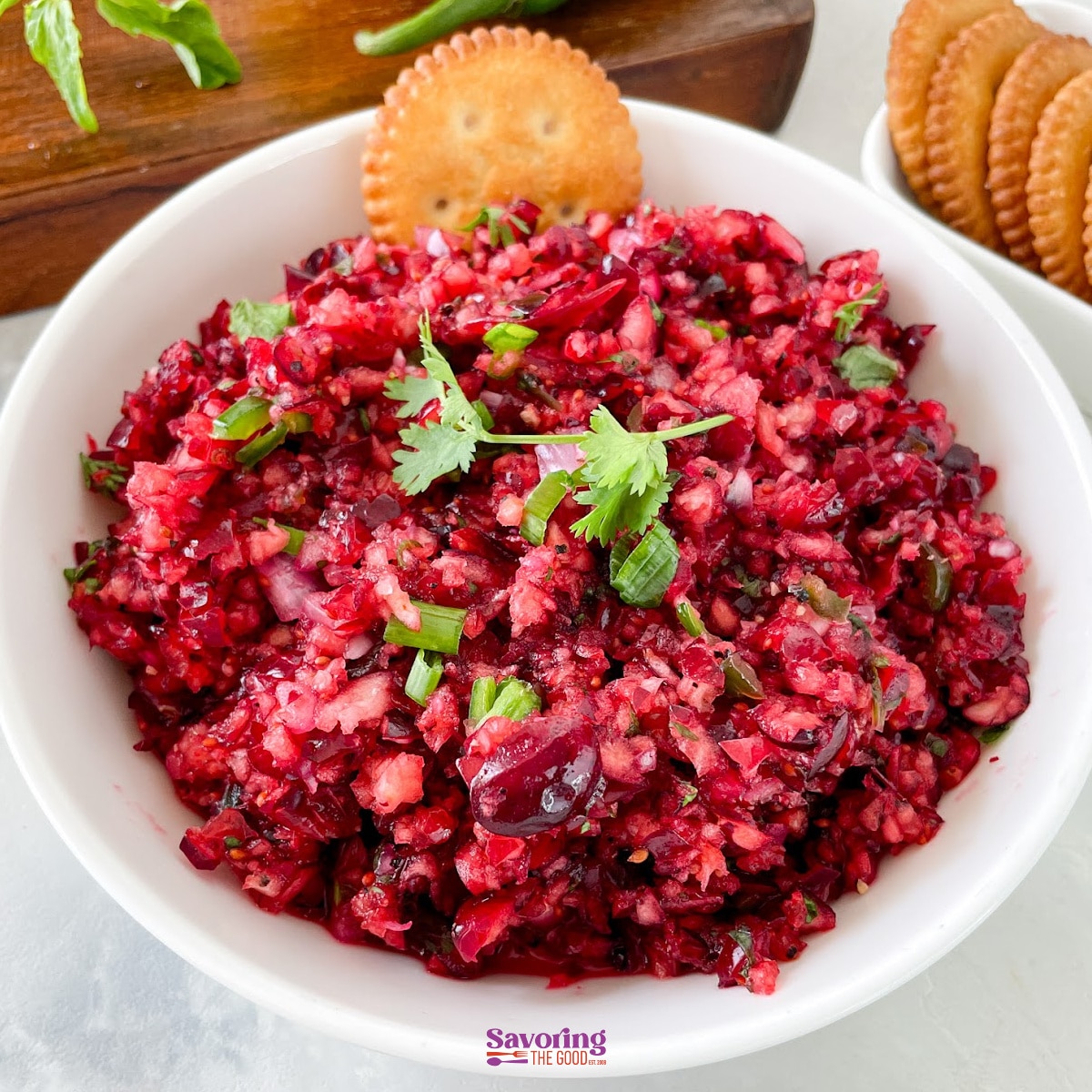 A white bowl filled with finely chopped cranberry salsa, garnished with cilantro, sits next to crackers on a white surface-a vibrant and refreshing twist for your next appetizer spread.