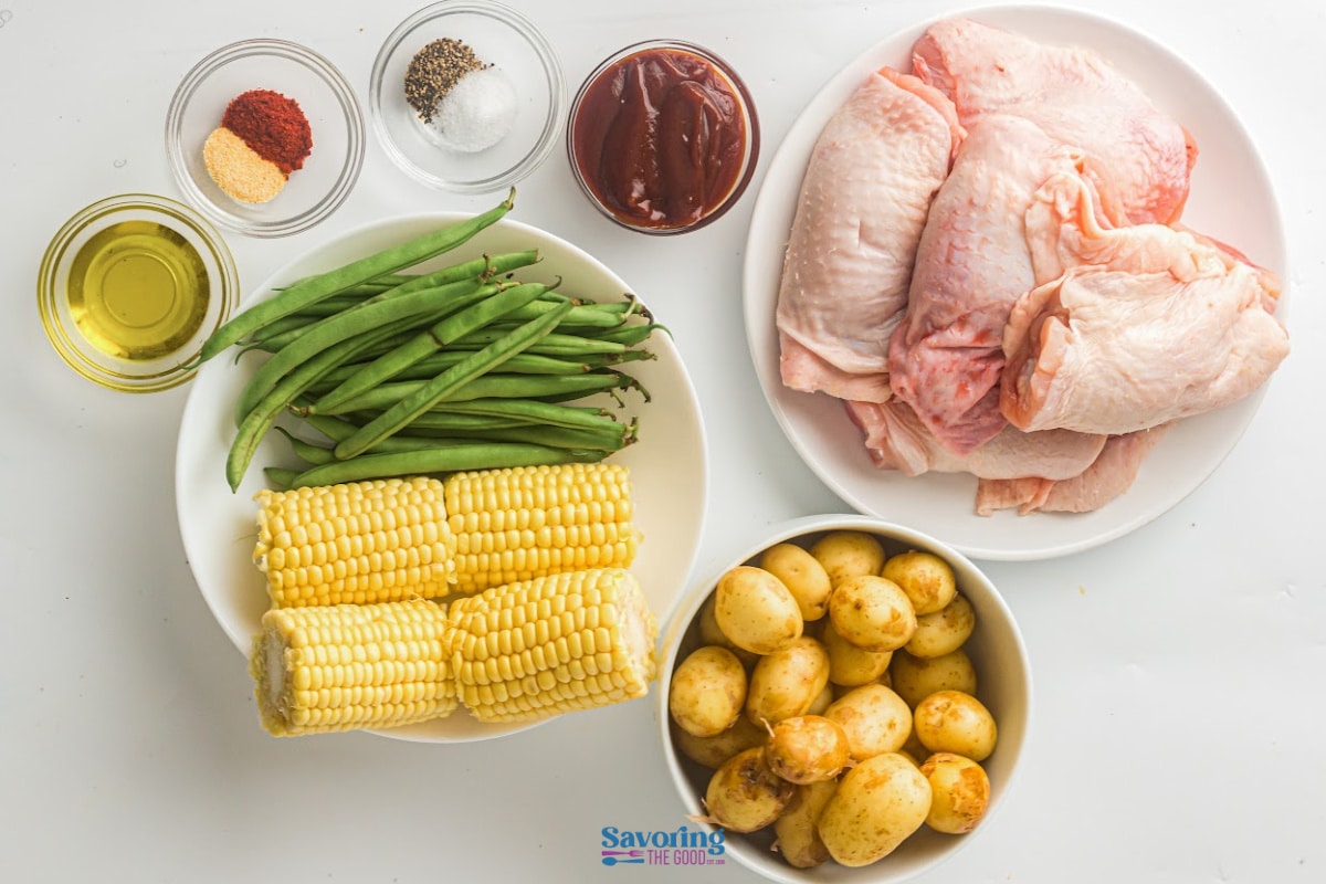 Raw ingredients on a white background: chicken pieces, baby potatoes, corn on the cob, green beans, olive oil, barbecue sauce, salt, pepper, paprika, and cayenne powder.