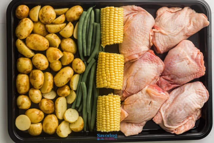 A baking tray with rows of halved baby potatoes, green beans, corn on the cob pieces, and raw chicken thighs.