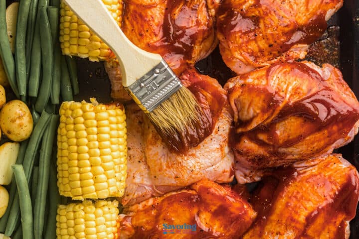 A brush applies barbecue sauce to raw chicken pieces on a tray, with corn on the cob, green beans, and small potatoes arranged beside the meat.