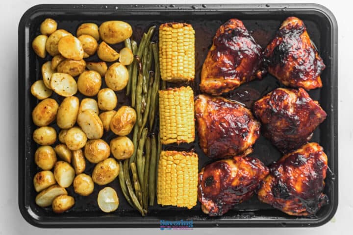 Sheet pan with roasted baby potatoes, green beans, corn on the cob, and barbecue-glazed chicken thighs arranged in sections.