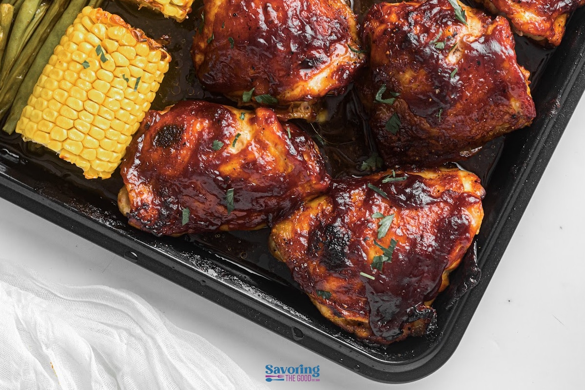 Baked barbecue chicken thighs on a tray with corn on the cob and green beans, garnished with herbs.