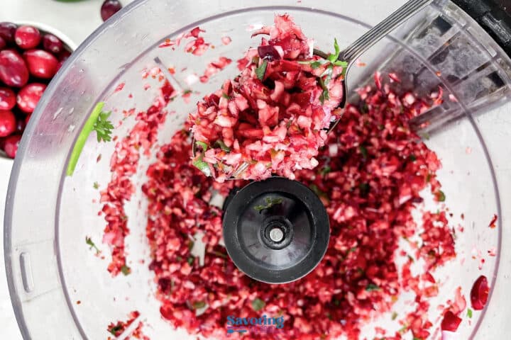 Chopped cranberry salsa mixture with herbs in a food processor, with a spoon scooping some out. Fresh cranberries are visible on the side.