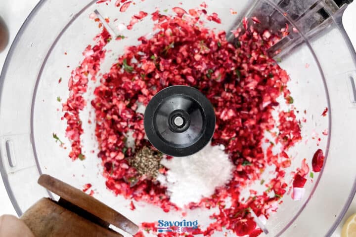 Chopped cranberries, sugar, and pepper are blended for a zesty cranberry salsa in a food processor, viewed from above as a hand adds freshly ground pepper.
