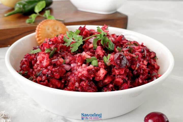 A white bowl filled with chunky cranberry salsa, garnished with cilantro, sits on a white surface next to a cracker.