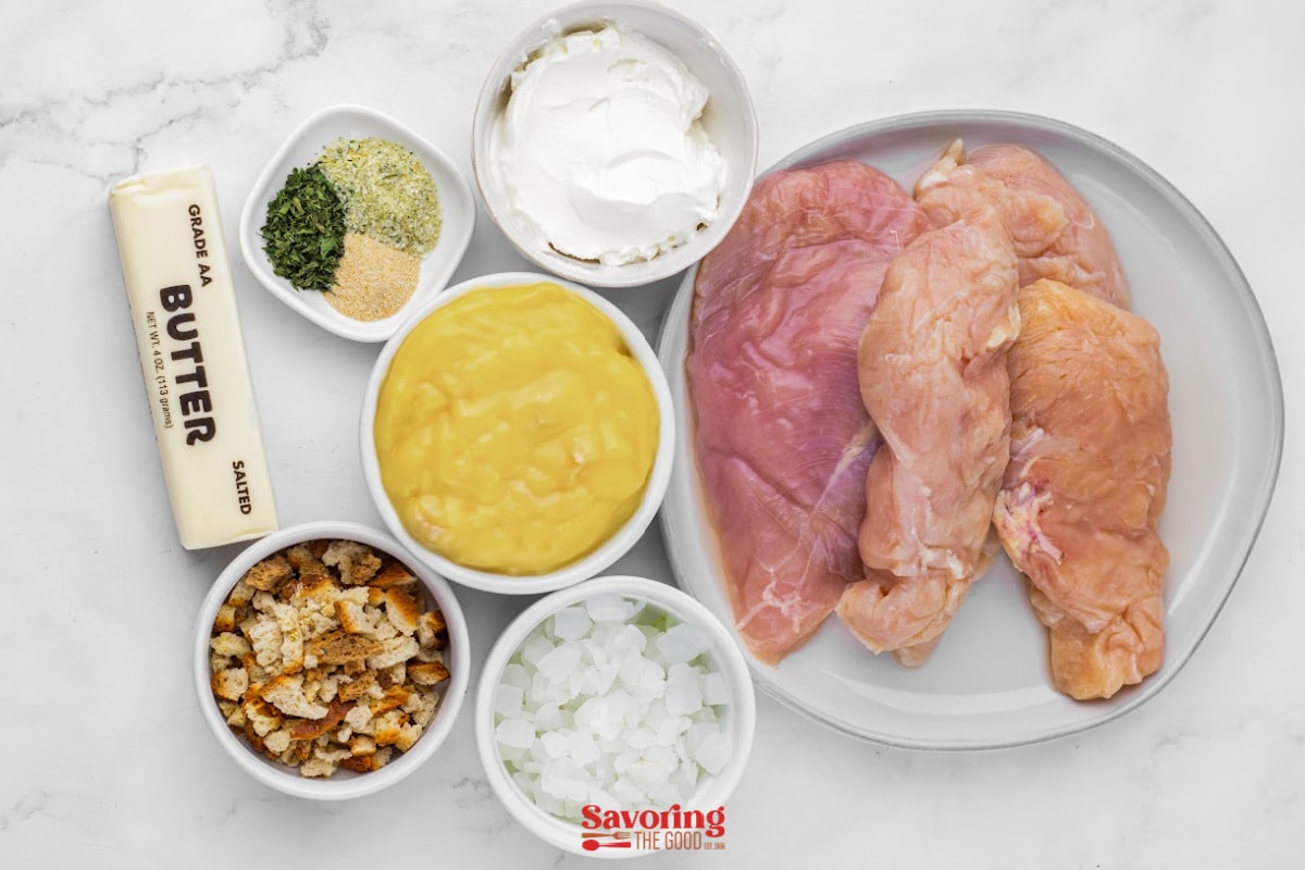 Raw chicken breasts on a plate surrounded by butter, seasoning, sour cream, cream of chicken soup, chopped onions, and stuffing mix-perfect for a comforting stuffing and chicken crock pot recipe.