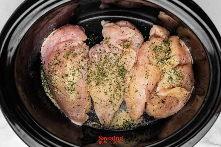 Three raw, seasoned chicken breasts placed inside a black slow cooker, sprinkled with herbs and spices.