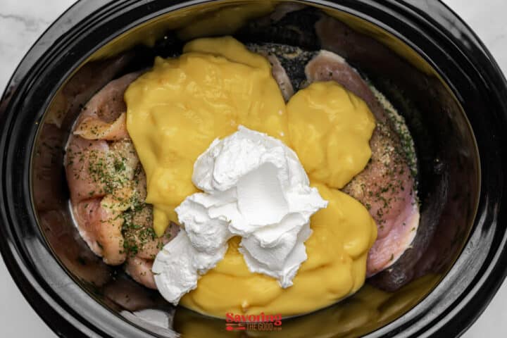 Raw chicken breasts in a slow cooker topped with cream of chicken soup, seasonings, and a dollop of cream cheese, ready to be cooked.