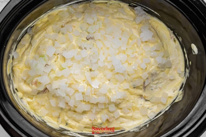 A mixture of creamy batter topped with diced white onions in a black slow cooker.