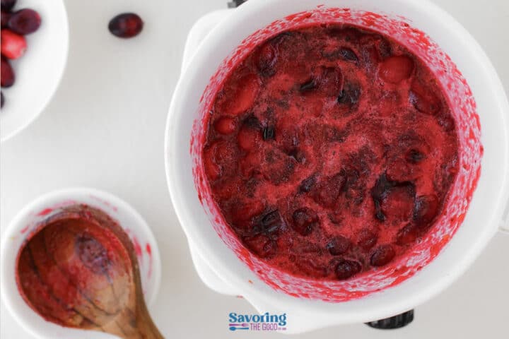 Overhead view of a white bowl filled with cooked cranberry sauce, next to a wooden spoon and some loose cranberries on a white surface.
