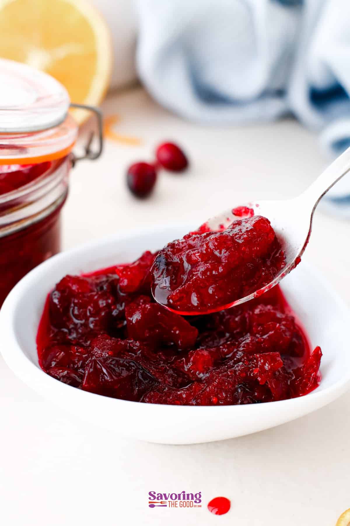 A spoonful of chunky cranberry sauce is held over a white bowl, with a jar of sauce, cranberries, and a lemon in the background.