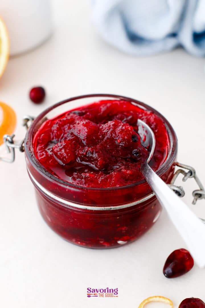 A glass jar filled with cranberry sauce sits on a white surface, with a spoon dipping into the sauce.