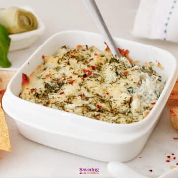 A white square dish filled with baked spinach and artichoke dip, topped with melted cheese and herbs, with a spoon inside.
