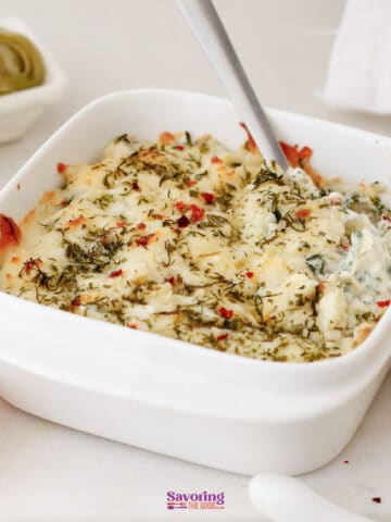 A white square dish filled with baked spinach and artichoke dip, topped with melted cheese and herbs, with a spoon inside.