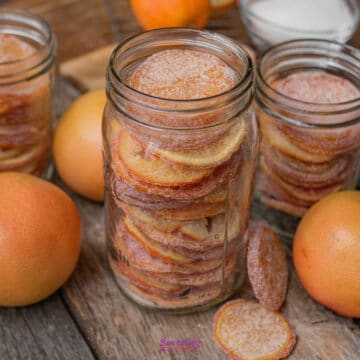 Glass jars filled with sugared citrus slices are surrounded by whole citrus fruits and a bowl of sugar on a wooden surface.