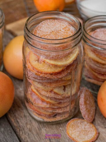 Glass jars filled with sugared citrus slices are surrounded by whole citrus fruits and a bowl of sugar on a wooden surface.