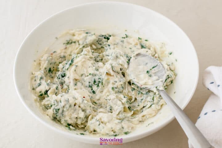 A white bowl filled with a creamy spinach artichoke dip mixture, with a metal spoon resting inside.