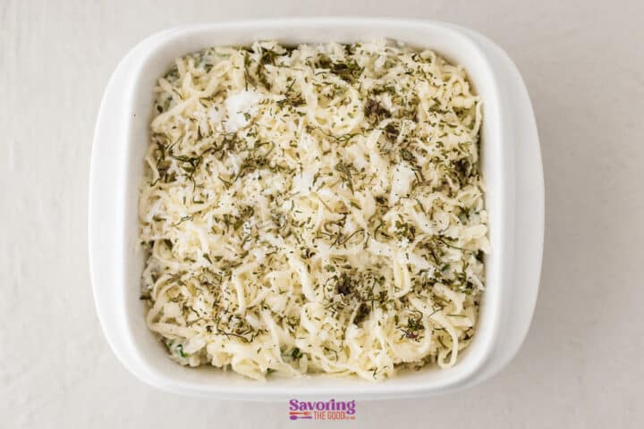 A white casserole dish filled with creamy spinach artichoke dip, topped with shredded cheese, herbs, and grated parmesan, ready to be baked.