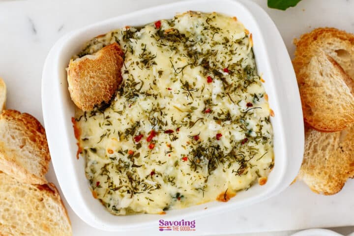 A white dish filled with creamy spinach artichoke dip, topped with melted cheese and herbs, served alongside slices of toasted bread.