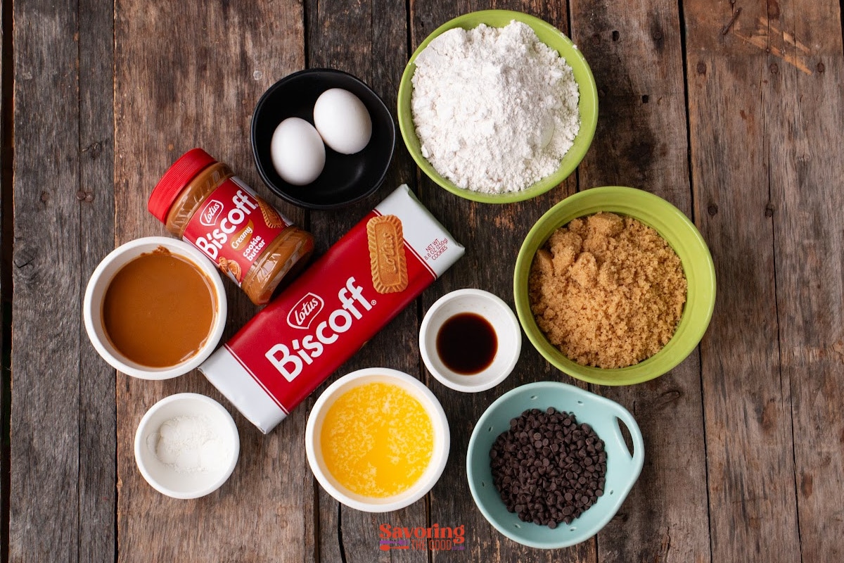 Ingredients for baking are arranged on a wooden surface, including eggs, Biscoff cookies and spread, flour, brown sugar, powdered sugar, vanilla, melted butter, chocolate chips, and baking powder-perfect for creating biscoff butter cookies.