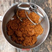 A metal mixing bowl with a paddle attachment contains a creamed brown sugar and biscoff butter cookie mixture on a rustic wooden surface.