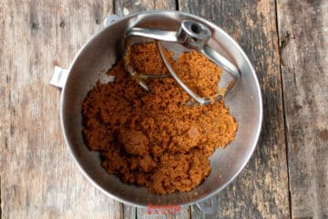 A metal mixing bowl with a paddle attachment contains a creamed brown sugar and biscoff butter cookie mixture on a rustic wooden surface.