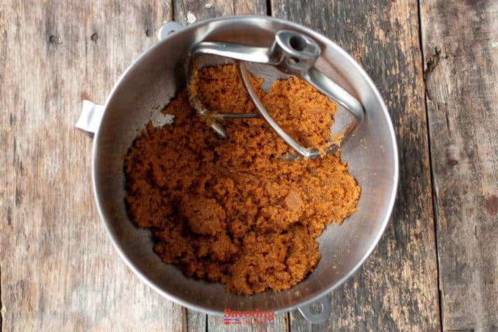 A metal mixing bowl with a paddle attachment contains a creamed brown sugar and biscoff butter cookie mixture on a rustic wooden surface.