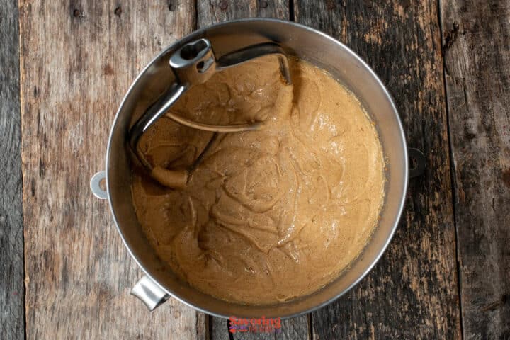 A mixing bowl with biscoff butter cookie batter and a metal beater attachment sits on a rustic wooden surface.