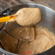 A hand holds a yellow spatula with thick, biscoff butter cookie batter above a metal mixing bowl containing more batter on a wooden surface.