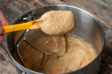 A hand holds a yellow spatula with thick, biscoff butter cookie batter above a metal mixing bowl containing more batter on a wooden surface.