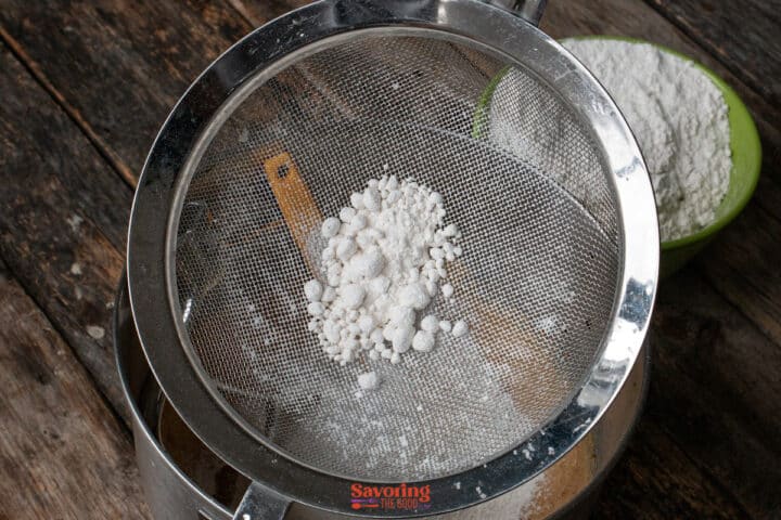 A metal sifter containing clumps of flour is held above a wooden surface, with a green bowl of flour nearby.