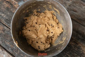 A metal mixing bowl filled with biscoff butter cookie dough sits on a rustic wooden table.