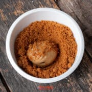 A scoop of ice cream sits in a white bowl filled with crumbled Biscoff butter cookie on a rustic wooden surface.