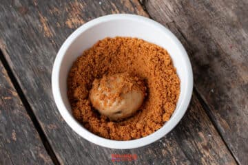 A scoop of ice cream sits in a white bowl filled with crumbled Biscoff butter cookie on a rustic wooden surface.