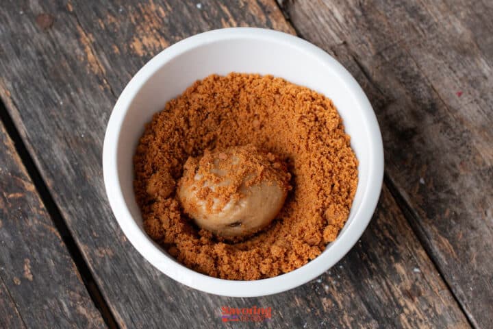 A scoop of ice cream sits in a white bowl filled with crumbled Biscoff butter cookie on a rustic wooden surface.