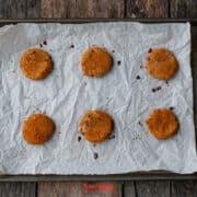 Six round biscoff butter cookies are spaced out on a parchment-lined baking sheet, resting on a rustic wooden surface.