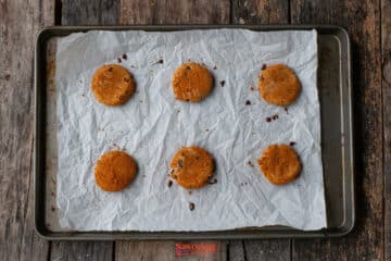 Six round biscoff butter cookies are spaced out on a parchment-lined baking sheet, resting on a rustic wooden surface.
