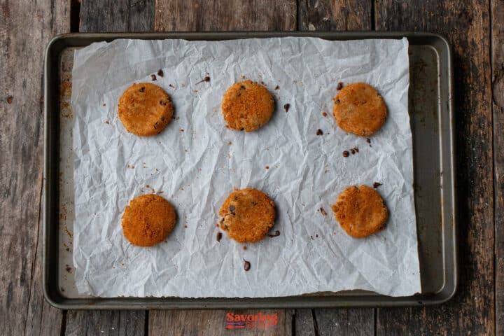 Six round biscoff butter cookies are spaced out on a parchment-lined baking sheet, resting on a rustic wooden surface.