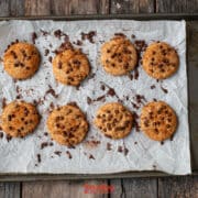 Eight chocolate chip and biscoff butter cookies are arranged on parchment paper atop a baking tray, with some crumbs scattered around, on a rustic wooden surface.
