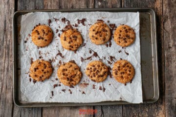 Eight chocolate chip and biscoff butter cookies are arranged on parchment paper atop a baking tray, with some crumbs scattered around, on a rustic wooden surface.
