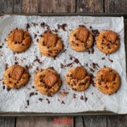 A baking tray on a wooden surface holds eight biscoff butter cookies topped with biscuits and chocolate chips on parchment paper.