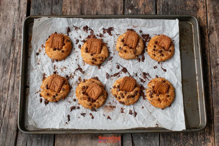 A baking tray on a wooden surface holds eight biscoff butter cookies topped with biscuits and chocolate chips on parchment paper.