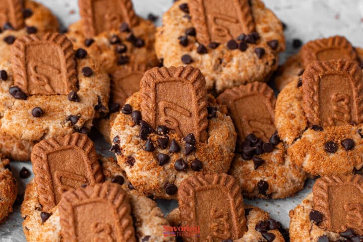 Cookies topped with chocolate chips and biscoff butter cookie pieces, arranged closely together on a light surface.