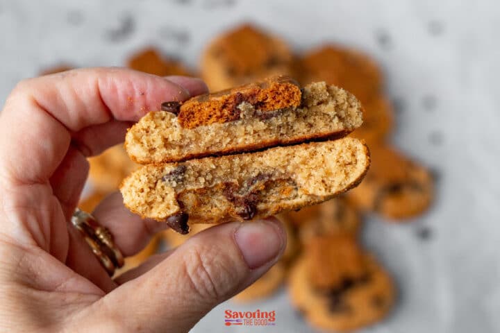 A hand holding two halves of a biscoff butter cookie, showing the soft cookie interior with chocolate chips. Other cookies are blurred in the background.