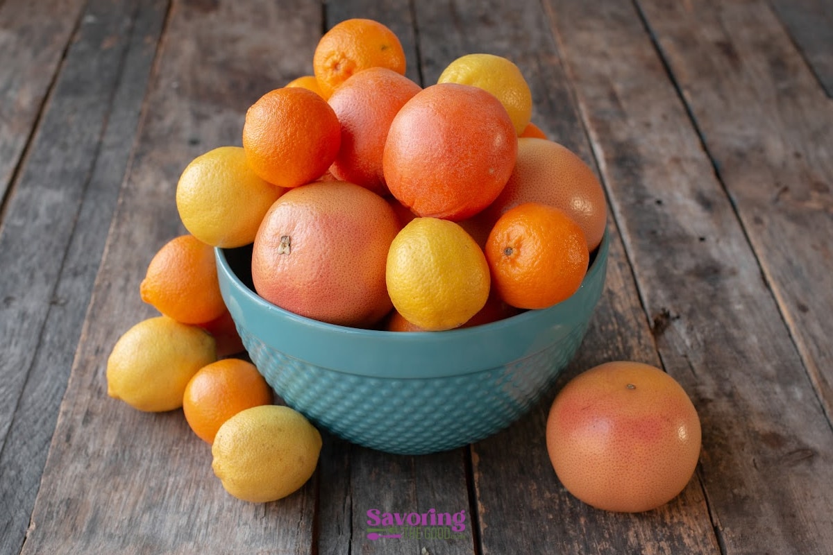 A blue bowl filled with assorted citrus fruits, including grapefruits, oranges, and lemons, sits on a wooden surface, with more citrus fruits scattered around it.