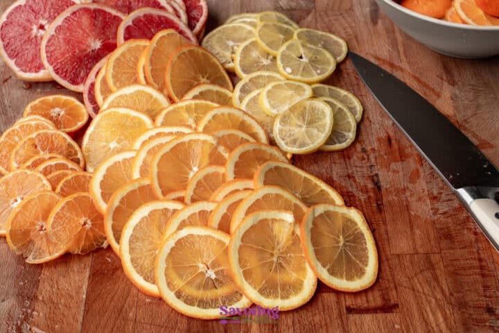 Thinly sliced oranges, lemons, limes, and grapefruit arranged on a wooden cutting board next to a kitchen knife.