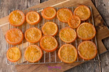 Slices of candied orange laid out in rows on a wire cooling rack placed over a wooden cutting board.