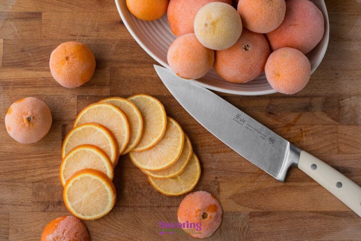 A knife rests on a wooden surface beside a bowl of whole oranges and several neatly sliced orange pieces.