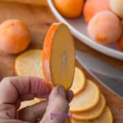A hand holds a thin slice of orange fruit above a cutting board with more slices, a knife, and a plate of whole fruits in the background.